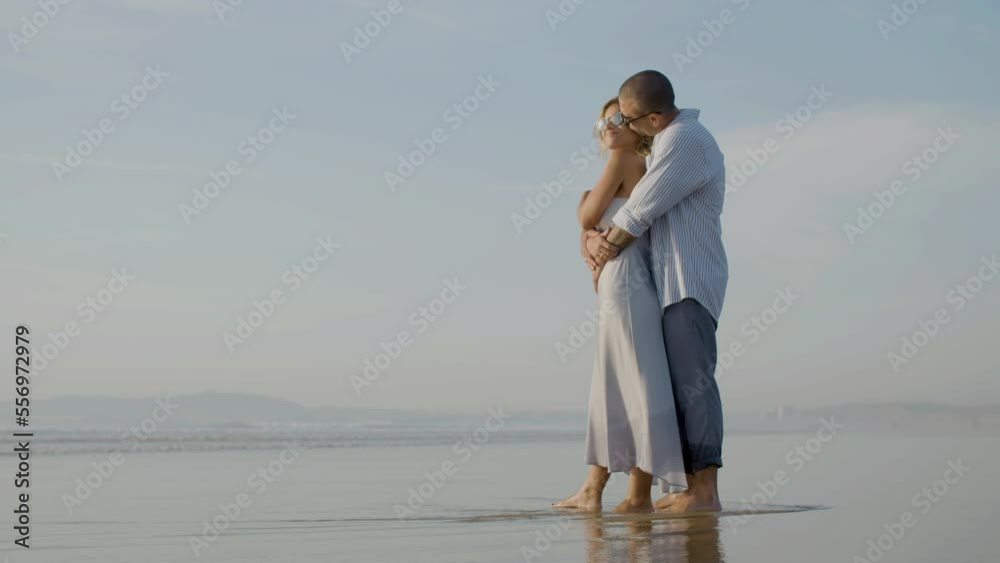 Happy Caucasian couple in sunglasses watching sunset at seashore. Middle-aged bearded man hugging beautiful blonde woman and kissing her head while enjoying precious moment together. Romance concept. Happy Caucasian couple in sunglasses watching sunset at seashore. Middle-aged bearded man hugging beautiful blonde woman and kissing her head while enjoying precious moment together. Romance concept.