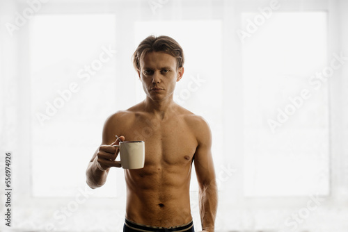 sporty man with a naked torso drinks from a cup in the morning by the window. © velimir