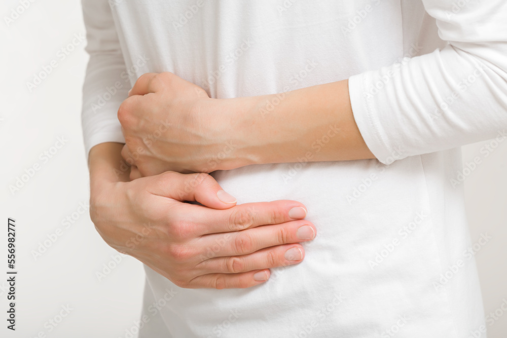 Young adult woman in white clothes touching painful belly with arms isolated on light gray background. Closeup. Side view. Girl suffering from stomach ache. Young adult woman in white clothes touching painful belly with arms isolated on light gray background. Closeup. Side view. Girl suffering from stomach ache.