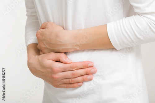 Young adult woman in white clothes touching painful belly with arms isolated on light gray background. Closeup. Side view. Girl suffering from stomach ache.