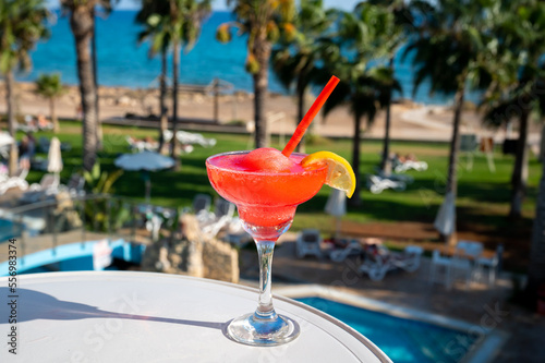 Colourful cold Strawberry daiquiri cocktail drink served in glass at pool bar overlooking blue pool, sea and palm trees, relax and holidays at sea