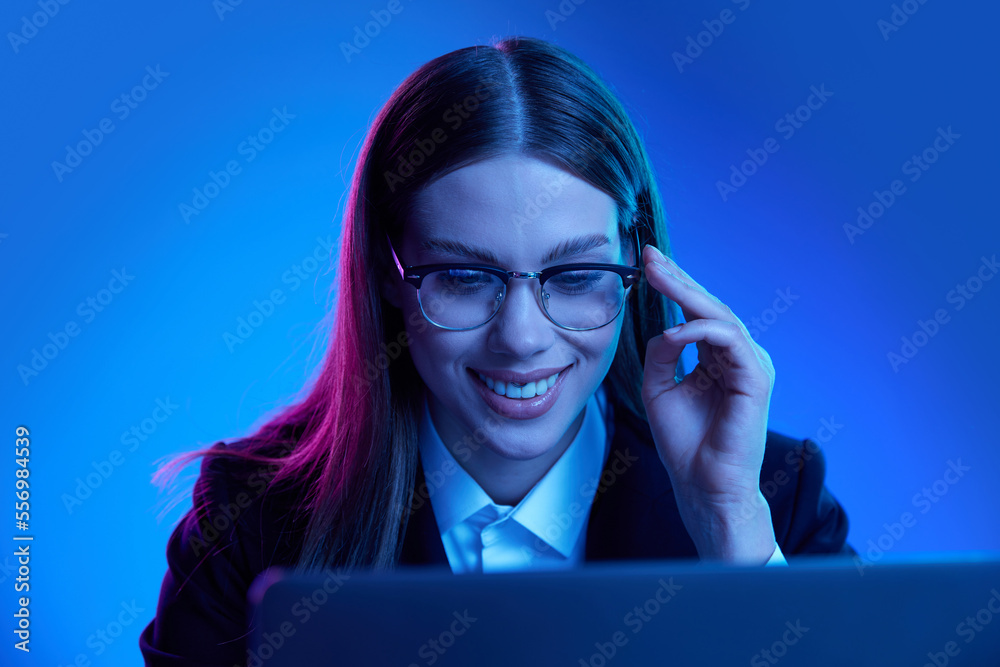 Woman in stylish official suit working on laptop over blue background in neon light. Marketing department Woman in stylish official suit working on laptop over blue background in neon light. Marketing department