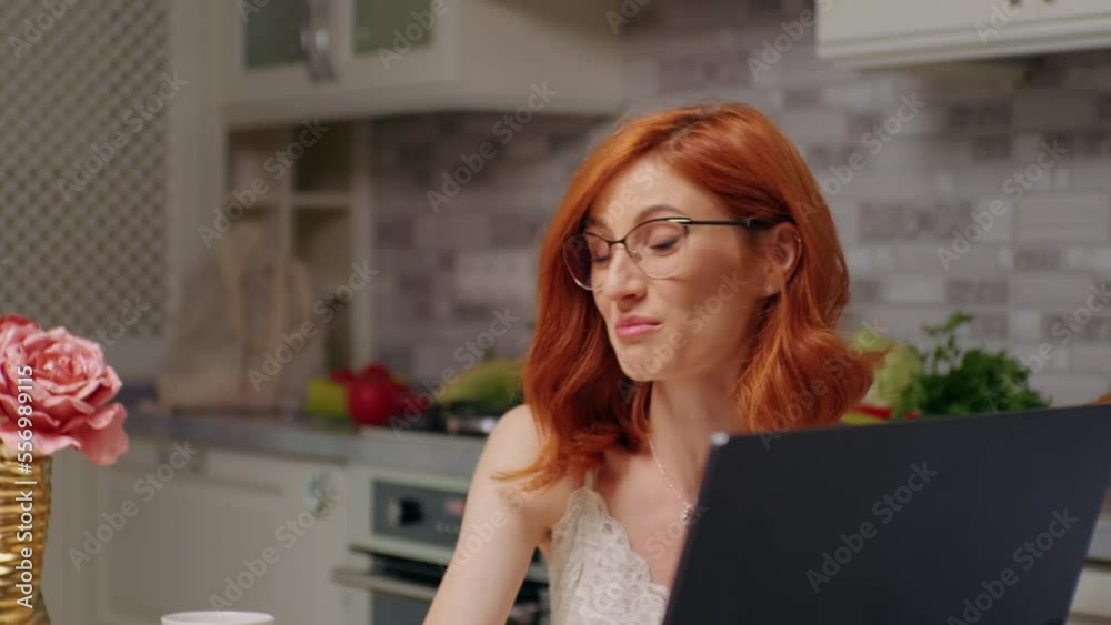 Female freelancer with red hair working on laptop in kitchen at table showing detailes to a friend. Small online business starting in personal kitchen. Female freelancer with red hair working on laptop in kitchen at table showing detailes to a friend. Small online business starting in personal kitchen.