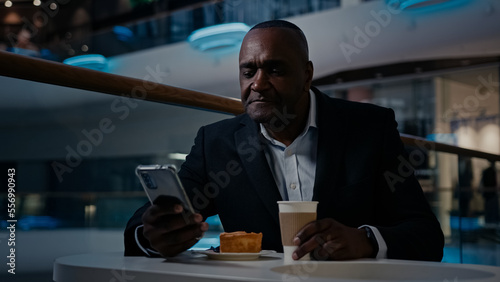 African adult middle-aged businessman 50s ethnic man in formal suit employer entrepreneur sitting at table in cafe drink coffee drinking tea browsing mobile phone smartphone app talk tell video call © Yuliia