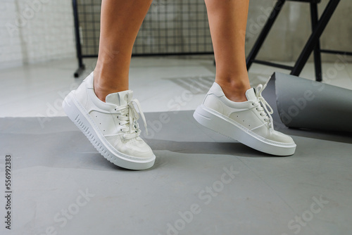 Beautiful woman in youth leather white sneakers and standing on white background. Women's legs with fashionable shoes. Casual white women's shoes
