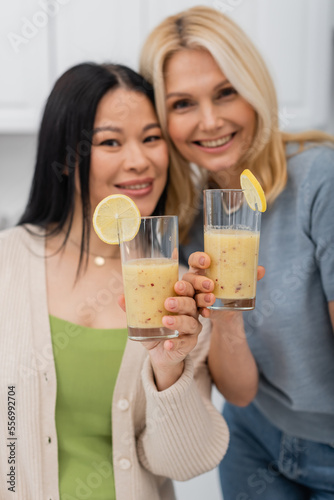 Blurred interracial friends looking at camera and holding fruit smoothie at home