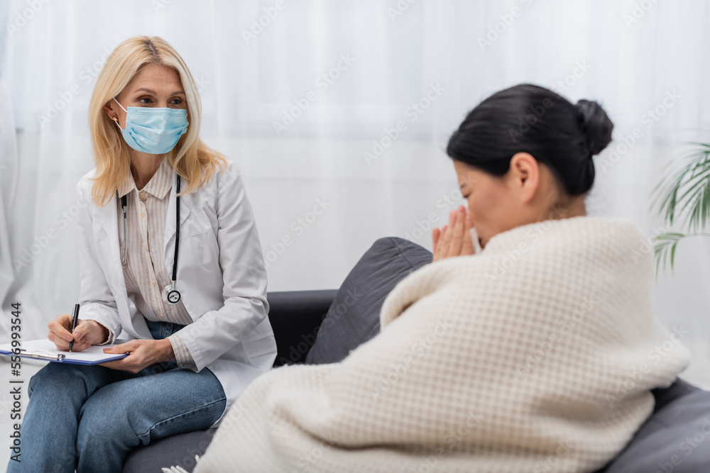 Doctor in medical mask holding clipboard near blurred asian patient at home