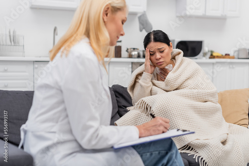 Diseased asian woman in blanket touching head near blurred doctor writing on clipboard at home