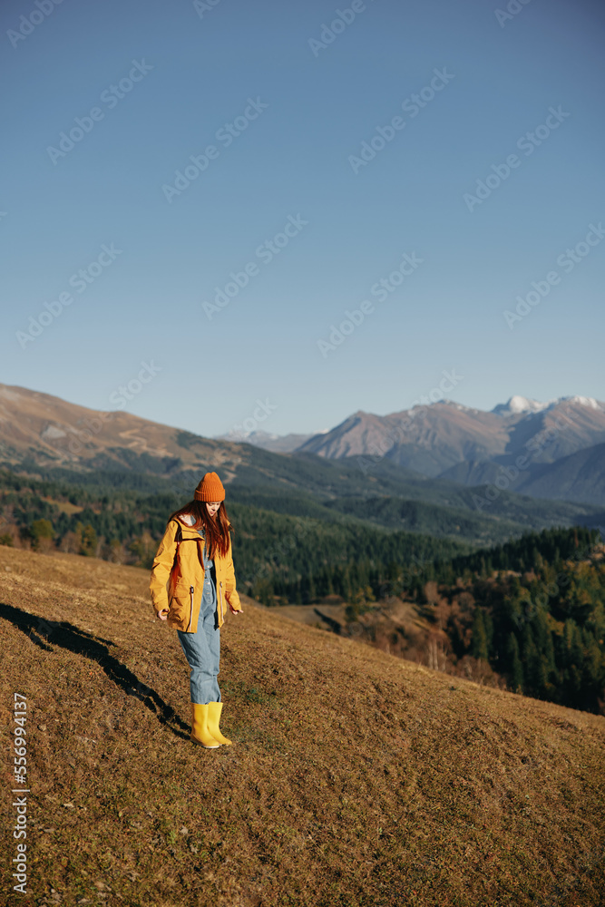 Full-length woman walking on a hill smiling with teeth and looking at the mountains in a yellow raincoat and jeans happy nature trip on a hike spring, freedom lifestyle 