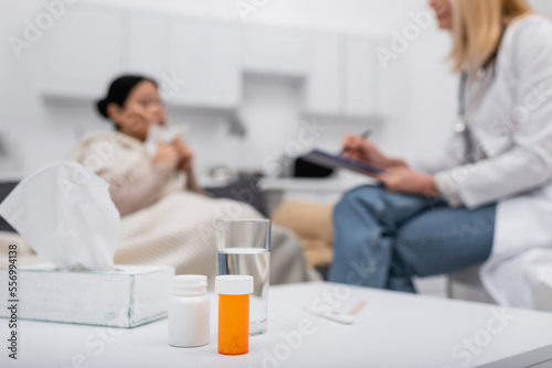 Pills and glass of water near blurred doctor and patient at home