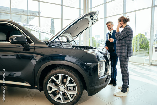 Businessmen standing near open front trunk thinking about buying car. Customer wearing costume holding tablet with documents of purchasing auto. Manager holding folder and selling automobile to client