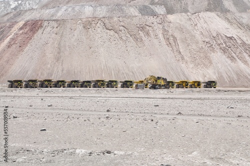 Huge mining trucks are parked in a row in front of a big hill. Mining trucks in Atacama desert, Chile. Lithium mining industry in Chile. Big mining trucks and diggers look like toys in the distance