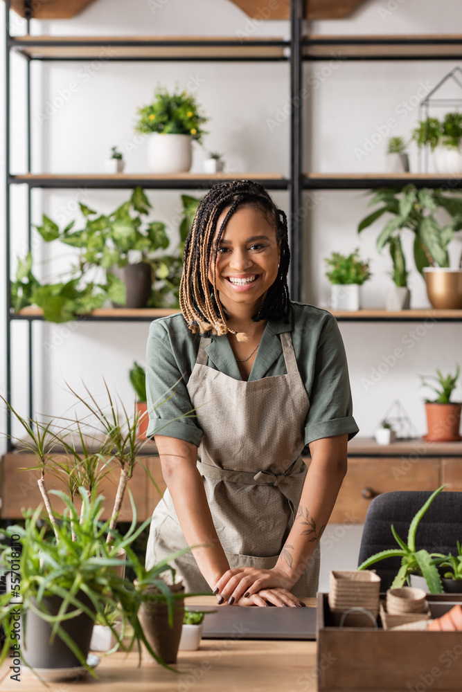 happy african american florist in apron and dreadlocks smiling at camera near laptop in flower shop happy african american florist in apron and dreadlocks smiling at camera near laptop in flower shop