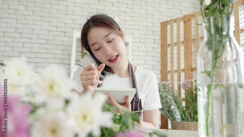 Smiling asian florist woman calling on smartphone and taking notes in notebook at flower shop, Female receiving order of flowers Smiling asian florist woman calling on smartphone and taking notes in notebook at flower shop, Female receiving order of flowers