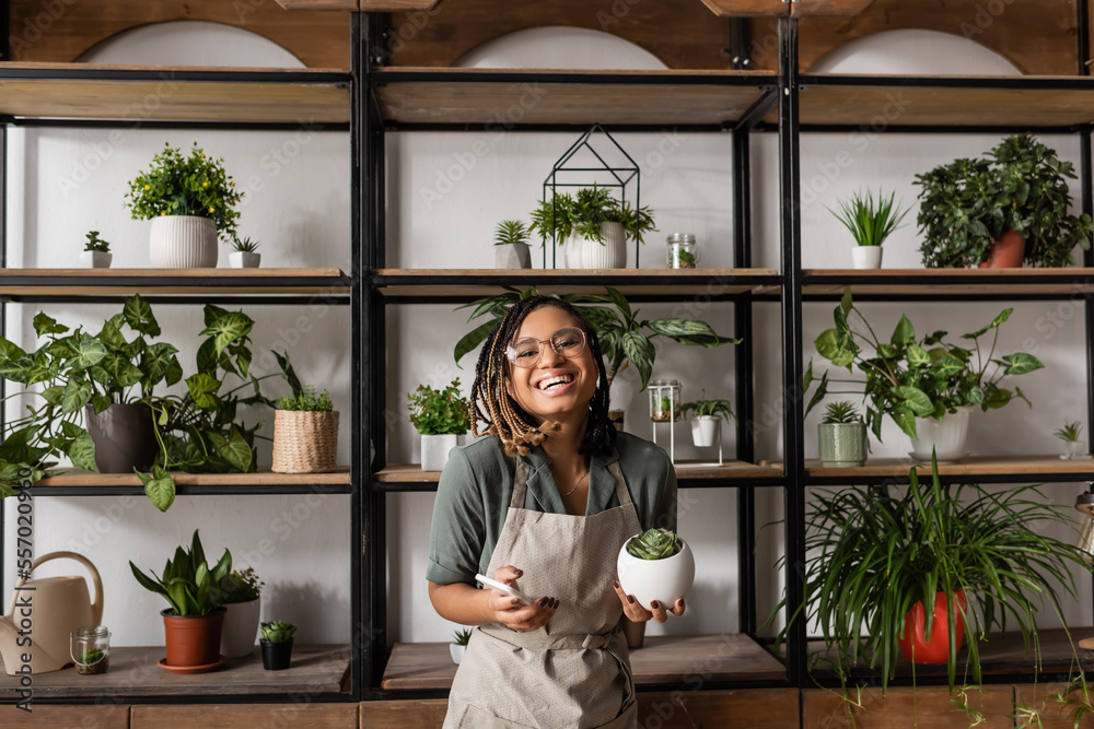 happy african american florist with mobile phone looking at camera near rack with different potted plants happy african american florist with mobile phone looking at camera near rack with different potted plants