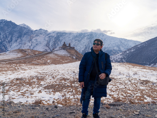 Asian tourist man in winter clothes standing in front of landscape view of Gergeti Holy Trinity Church on hill and Caucasus mountains - Tsminda Sameba. Beautiful Georgian landmark in early winter.   © Thitiporn