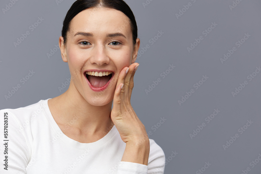 a happy woman stands on a gray background in a white tight-fitting T-shirt and smiling happily looks at the camera touching her neck with her hands
