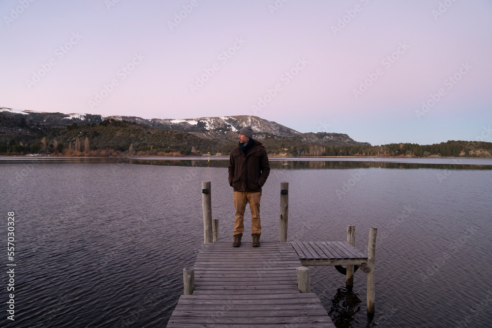 The placid lake at nightfall. Magical view of a man standing in the wooden dock with the mountains and forest in the background. Beautiful sunset colors in the sky and water.