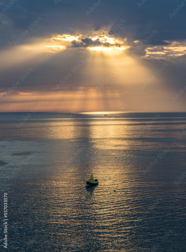 A lone boat sits on a calm sea, as crepuscular sun rays break through from behind the clouds at sunrise