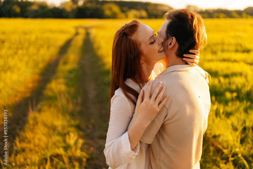 Smiling redhead young woman with closed eyes and unrecognizable man in love hugging, kissing, stroking standing together on beautiful green meadow, in summer evening during golden sunset. Smiling redhead young woman with closed eyes and unrecognizable man in love hugging, kissing, stroking standing together on beautiful green meadow, in summer evening during golden sunset.