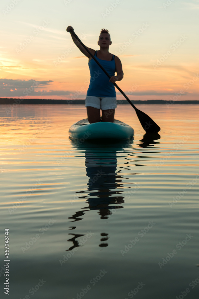 A woman with a mohawk in shorts on her knees on a sapboard with an oar against the backdrop of a sunset sky swims in the lake in the evening.