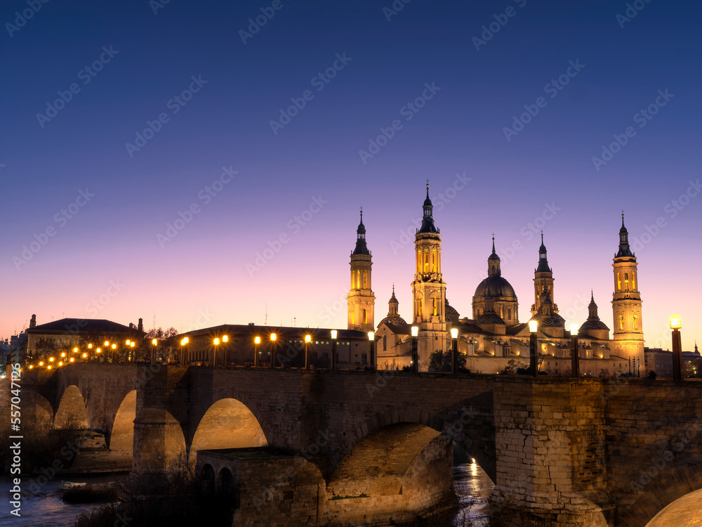 View of the basilica del Pilar de Zaragoza with the medieval stone bridge over the Ebro river in the foreground during sunset in winter.