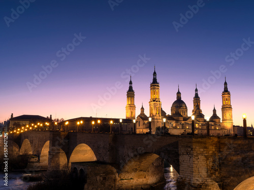 View of the basilica del Pilar de Zaragoza with the medieval stone bridge over the Ebro river in the foreground during sunset in winter.