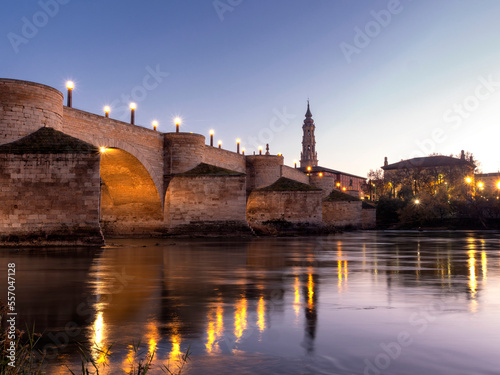 View of the tower of La Seo de Zaragoza cathedral with the medieval stone bridge over the Ebro river in the foreground during sunset in winter.