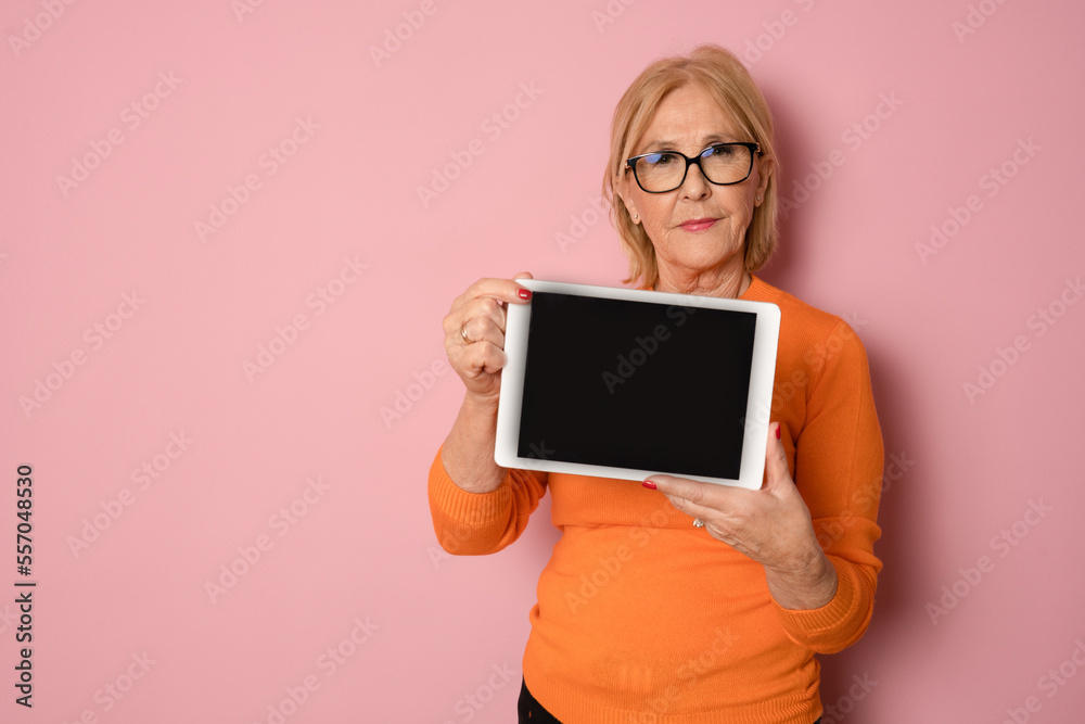 Senior woman holding black tablet isolated over pink background.