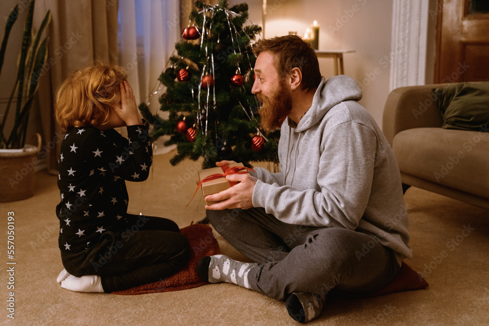 Boy covering eyes with hands while receiving Christmas gift from his father in cozy living room Boy covering eyes with hands while receiving Christmas gift from his father in cozy living room