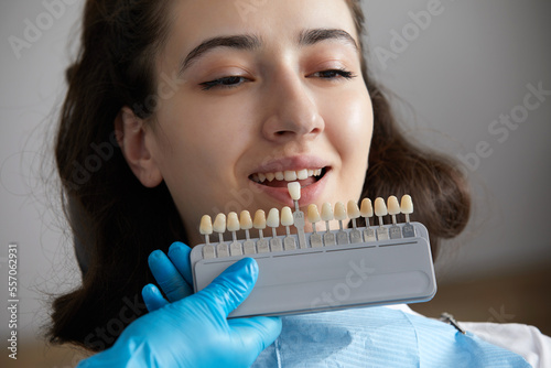 Dentist choosing color of tooth enamel for patient. Dentist applying sample from tooth enamel scale to caucasian female teeth