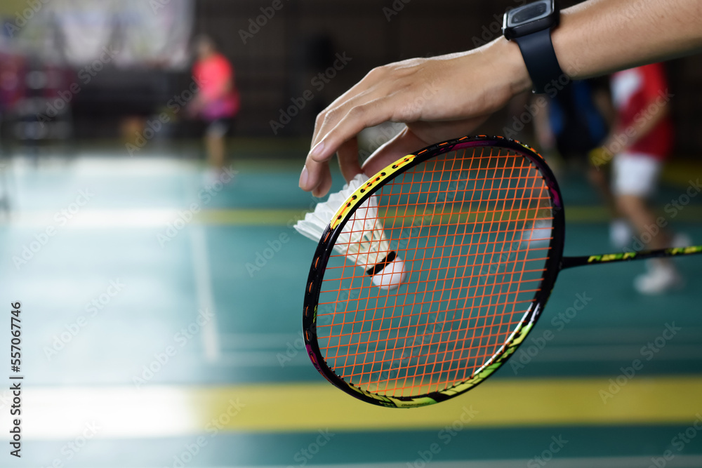 Badminton racket and old white shuttlecock holding in hands of player while serving it over the net ahead, blur badminton court background and selective focus.