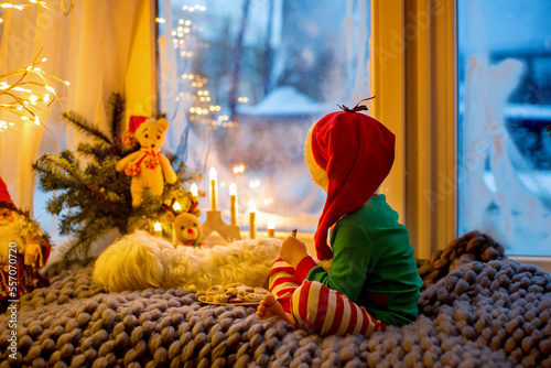 Toddler child, cute blond boy, sitting on the window, reading book, christmas lights around