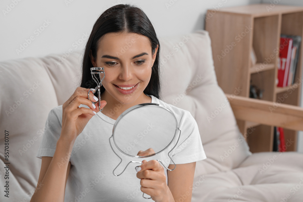 Beautiful young woman with mirror using eyelash curler indoors Beautiful young woman with mirror using eyelash curler indoors