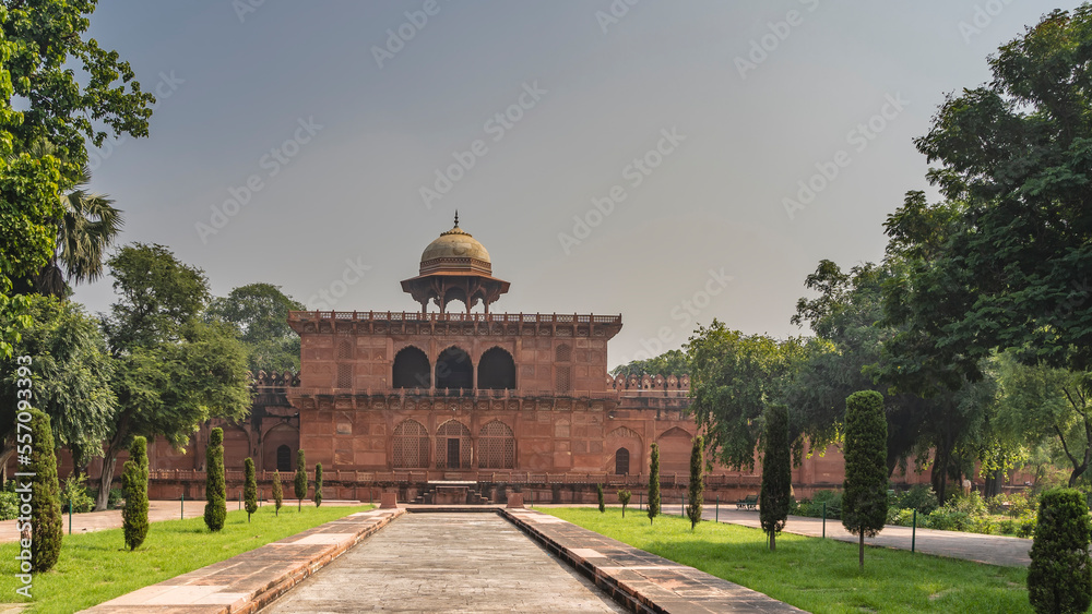 The territory of the ancient Taj Mahal complex. The building is built of red sandstone with arches, latticed windows, a dome with a spire. Trimmed bushes grow on the sides of the paved pedestrian path