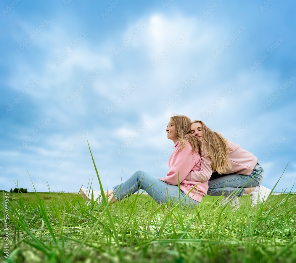 Two young girls are resting, relaxing, sitting on the grass by the sea. Hugging and smiling. Comfortable, casual clothes. Summer day. Two young girls are resting, relaxing, sitting on the grass by the sea. Hugging and smiling. Comfortable, casual clothes. Summer day.