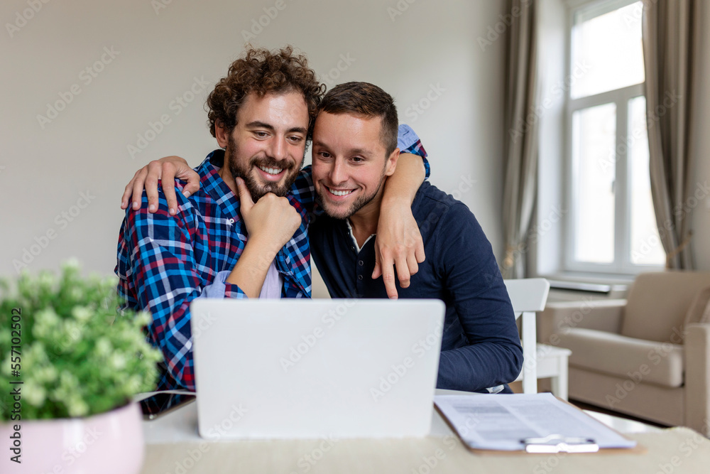 Lovely gay couple laughing together while sitting in their living room at home. Two romantic young male lovers having fun surfing the internet indoors. Young gay couple living together. Lovely gay couple laughing together while sitting in their living room at home. Two romantic young male lovers having fun surfing the internet indoors. Young gay couple living together.