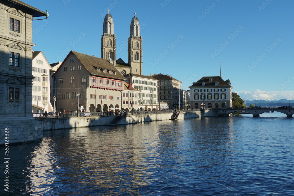 View on Limmat River bank surrounded by historical buildings enlighten by sun. In background is Quay Bridge in Zurich in front view and Grossmunster cathedral. 