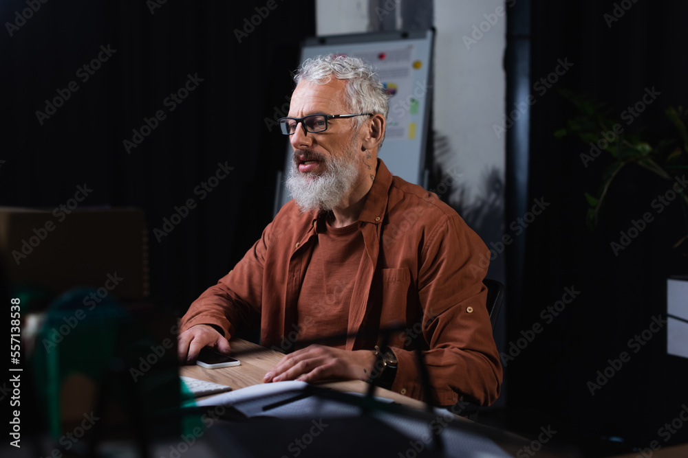 Fototapeta premium bearded businessman in eyeglasses holding smartphone while working in office at night