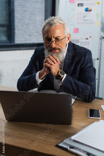 Focused mature businessman looking at laptop near papers and smartphone in office