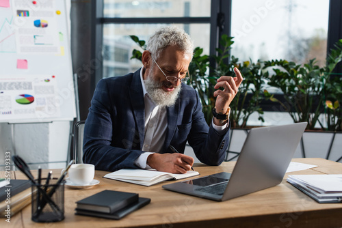 Middle aged businessman writing on notebook near laptop and coffee in office