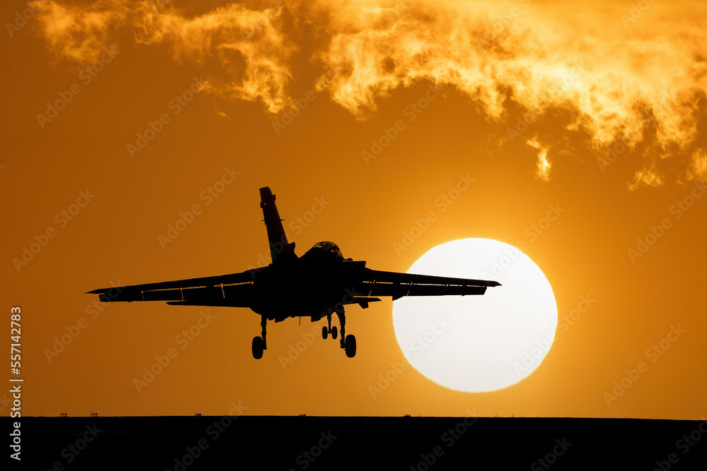 Fighter jet at sunset with orange sky and large setting sun after a combat mission. Flying over dramatic silhouette of fighter bomber jet aircraft - obrazy, fototapety, plakaty 