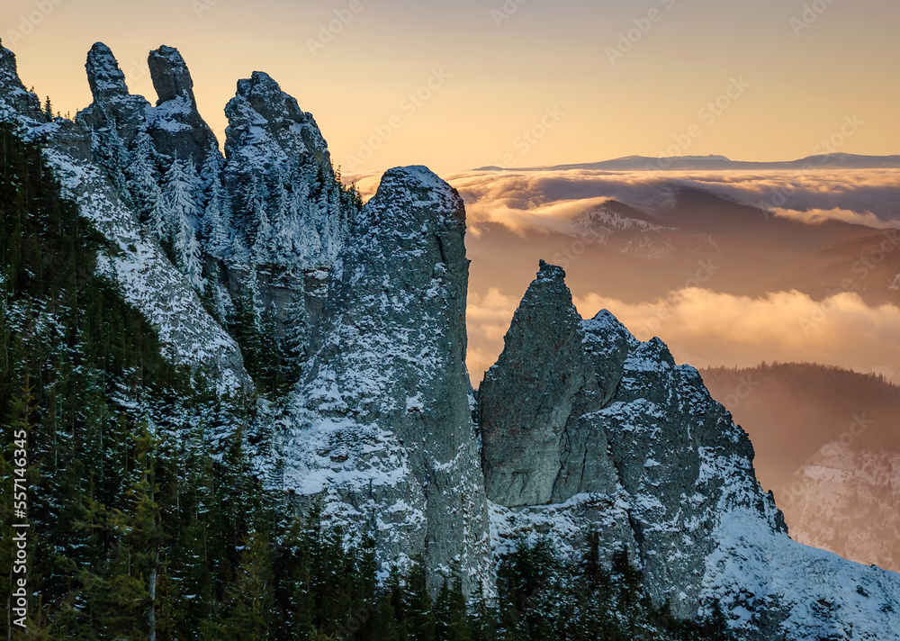 Dramatic evening winter landscape. Erroded mountain peaks covered in snow rise high in the foreground, background with foggy valleys and fluffy clouds. Aerial sunset view of snow covered mountains