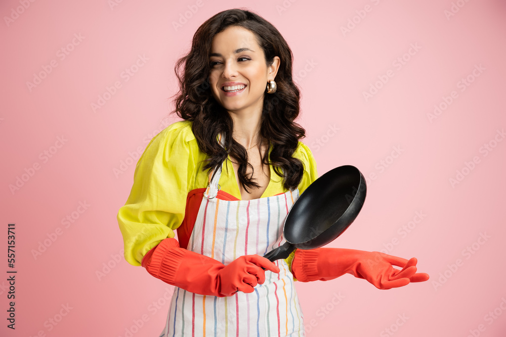 smiling brunette woman in yellow blouse and red rubber gloves holding frying pan while looking away isolated on pink smiling brunette woman in yellow blouse and red rubber gloves holding frying pan while looking away isolated on pink