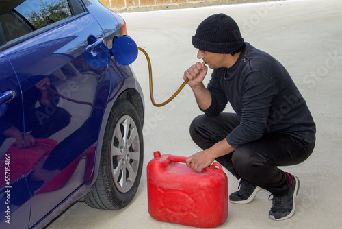 Obraz na plátně Image of a thief dressed in black who sucks petrol from a car tank with a rubber hose