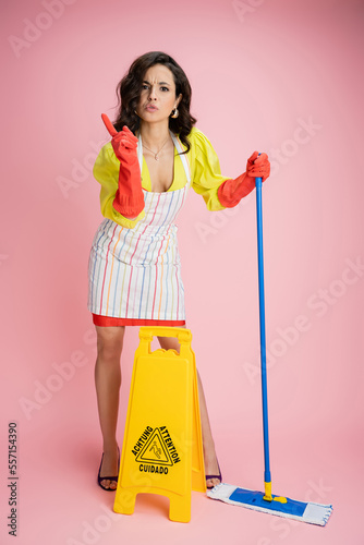 full length of strict housewife with mop wagging finger near wet floor caution sign on pink background
