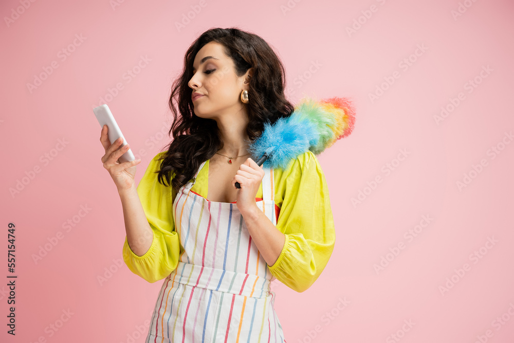 displeased housewife in striped apron holding colorful dust brush and looking at mobile phone isolated on pink displeased housewife in striped apron holding colorful dust brush and looking at mobile phone isolated on pink