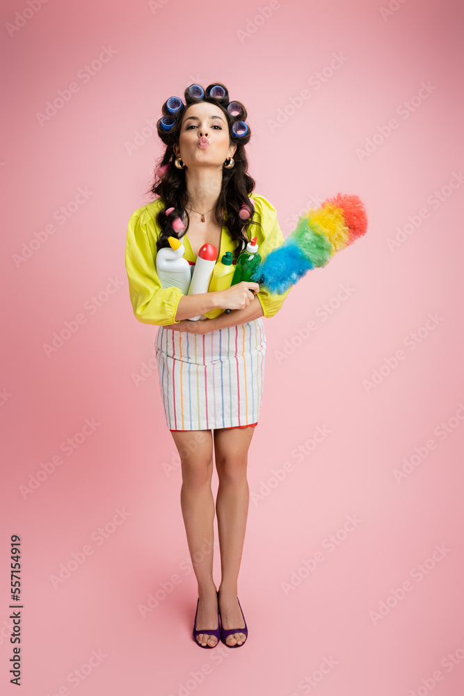 full length of flirty housewife in hair curlers holding dust brush and detergents while pouting lips on pink background