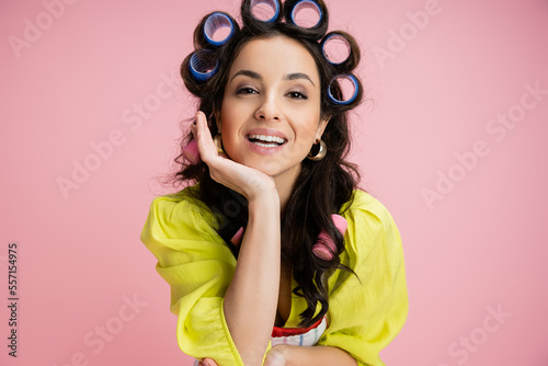 portrait of brunette woman in yellow blouse and hair curlers holding hand near face and looking at camera isolated on pink