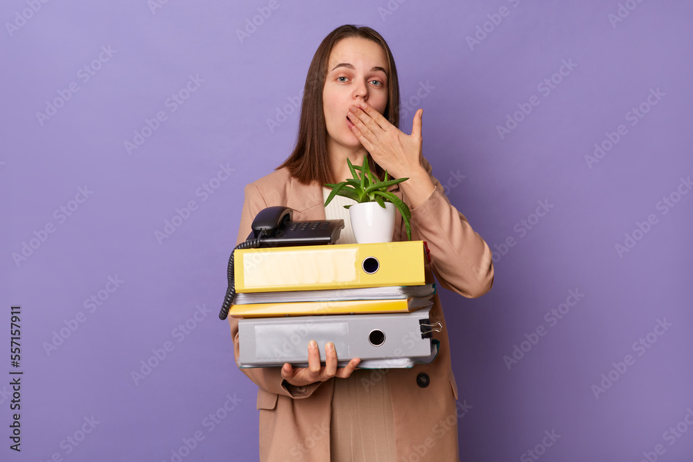 Portrait of shocked surprised woman wearing beige jacket holding lot of documents folders isolated over purple background, looking at camera, covering mouth with palm, having mistake at her work.